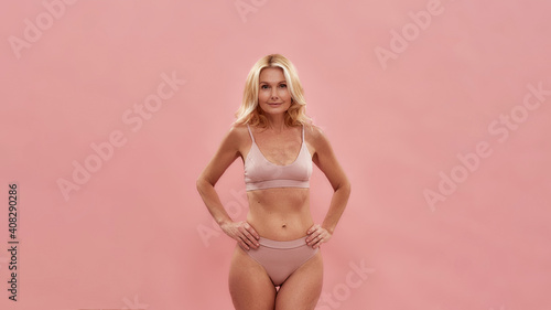 Beautiful mature woman wearing lingerie posing in studio. Happy middle aged caucasian woman in underwear smiling at camera while standing against pink background photo
