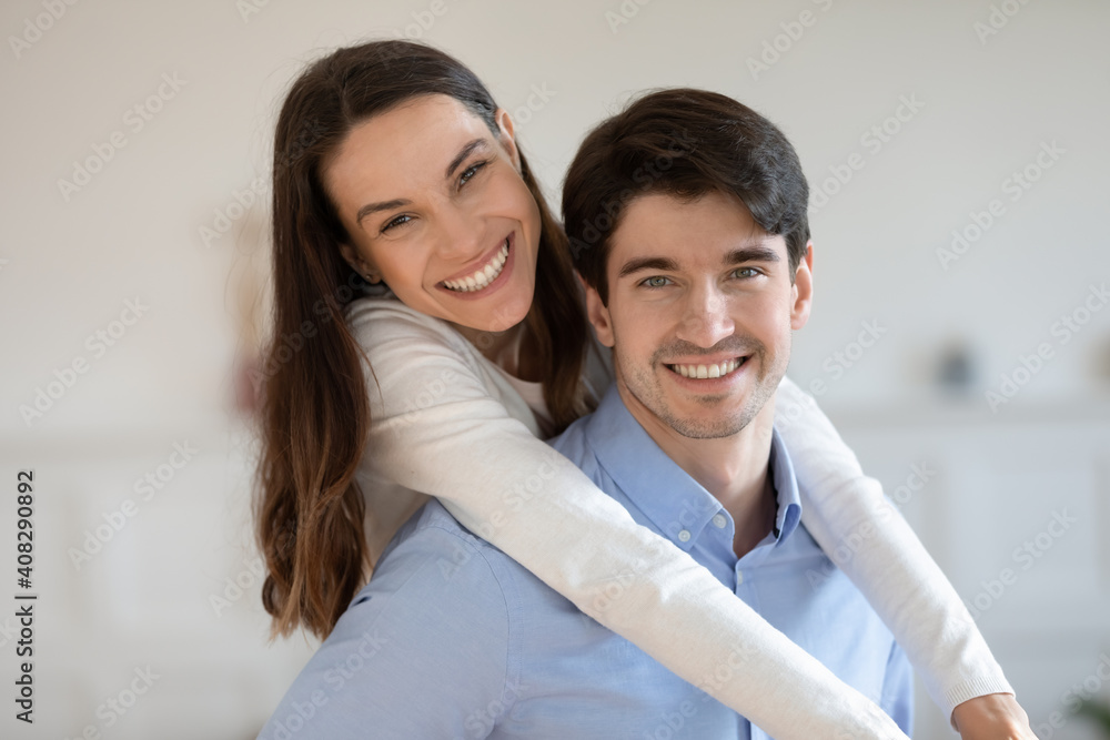 Head shot portrait smiling man piggy backing attractive woman, posing for funny family photo, standing at home, young couple enjoying weekend, happy wife and husband hugging, looking at camera Head shot portrait smiling man piggy backing attractive woman, posing for funny family photo, standing at home, young couple enjoying weekend, happy wife and husband hugging, looking at camera