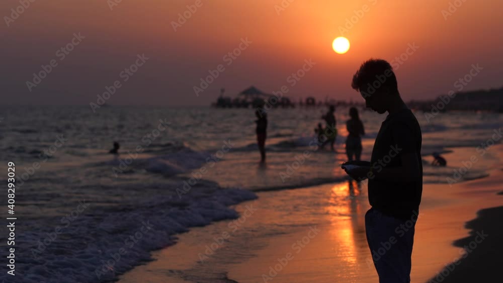 Black silhouette of handsome young kid standing alone on sunset sea beach. Boy puts medical face mask on and walks away Black silhouette of handsome young kid standing alone on sunset sea beach. Boy puts medical face mask on and walks away