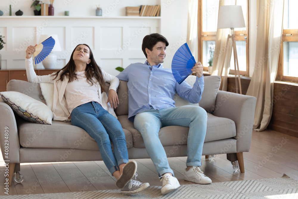 Exhausted man and woman waving blue paper fans, breathing, resting sitting on couch in living room, overheated tired couple suffering from hot summer weather, heating at home, feeling discomfort Exhausted man and woman waving blue paper fans, breathing, resting sitting on couch in living room, overheated tired couple suffering from hot summer weather, heating at home, feeling discomfort