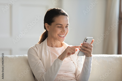 Close up overjoyed young woman looking at phone screen, sitting on cozy couch at home, smiling female reading message, chatting in social network or shopping, enjoying weekend with mobile device