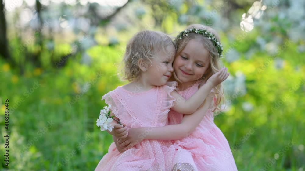 Two cute little girls sisters in pink dresses in a blooming spring park tenderly hug. A couple of happy children. happy family kid dream concept. Slow motion. Two cute little girls sisters in pink dresses in a blooming spring park tenderly hug. A couple of happy children. happy family kid dream concept. Slow motion.