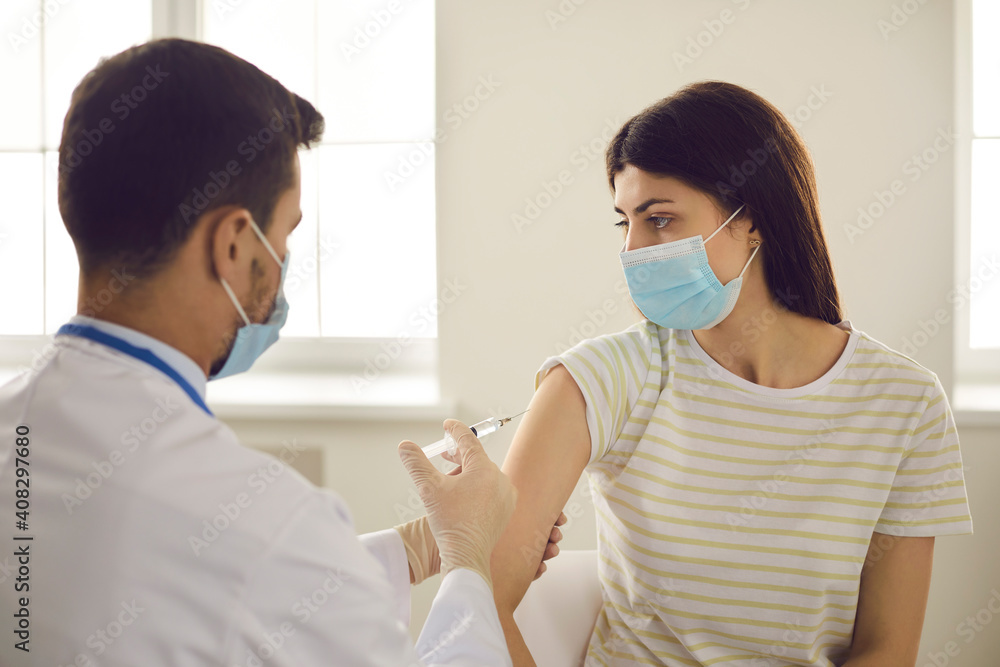 Doctor giving flu shot to female patient during seasonal immunization campaign at hospital. Young woman in medical face mask getting modern Covid-19 vaccine injection at clinic or health center