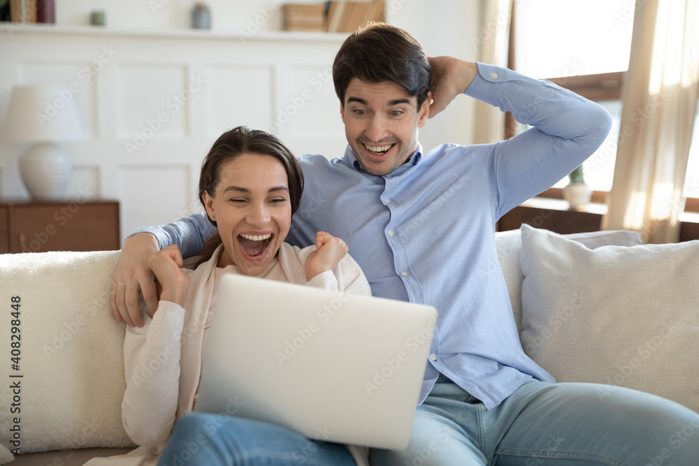 Close up overjoyed millennial couple looking at laptop screen, hugging, sitting on couch at home, happy woman and man reading great news, celebrating online lottery win, unexpected message Close up overjoyed millennial couple looking at laptop screen, hugging, sitting on couch at home, happy woman and man reading great news, celebrating online lottery win, unexpected message