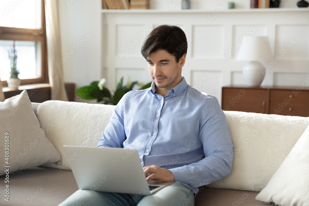 Focused man using laptop, sitting on cozy couch at home, looking at computer screen, chatting or shopping in internet, reading message, freelancer working online, spending leisure time with device Focused man using laptop, sitting on cozy couch at home, looking at computer screen, chatting or shopping in internet, reading message, freelancer working online, spending leisure time with device