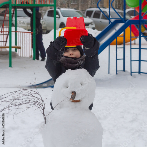 A preschool boy made a snowman out of snow in winter and puts a red bucket on the snowman's head