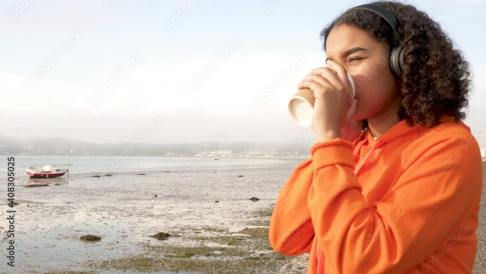 Beautiful mixed race African American girl teenager young woman wearing orange hoodie and wireless headphones, drinking takeout coffee by a harbor looking sad or thoughtful Beautiful mixed race African American girl teenager young woman wearing orange hoodie and wireless headphones, drinking takeout coffee by a harbor looking sad or thoughtful