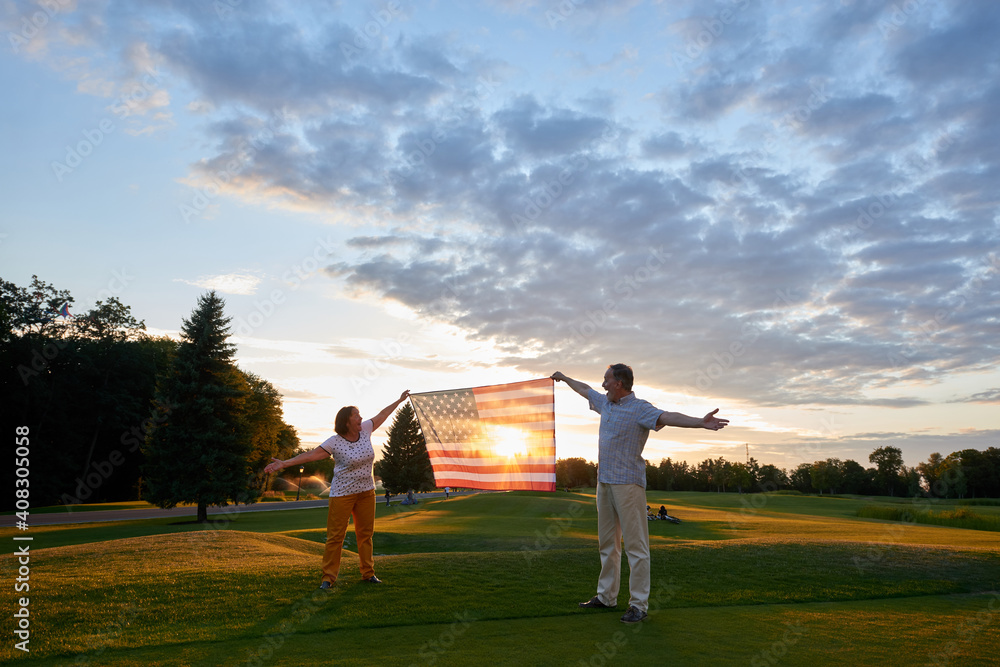 Mature couple holding the flag of the United States. Evening sunset sky and green field with short grass background. Mature couple holding the flag of the United States. Evening sunset sky and green field with short grass background.