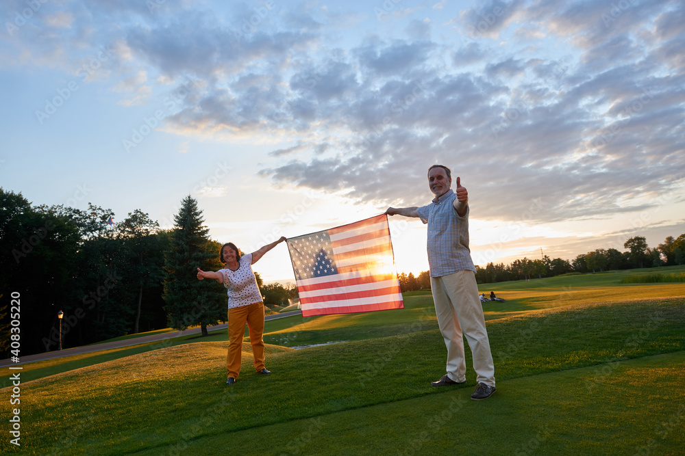 Obraz premium Couple holding us flag and showing thumb up. Patriotic man and woman on the green field.