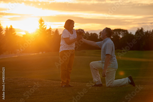 Retired man kneeking and giving flowers to his wife. Gifting bouquet outdoors against bright evening sunset.