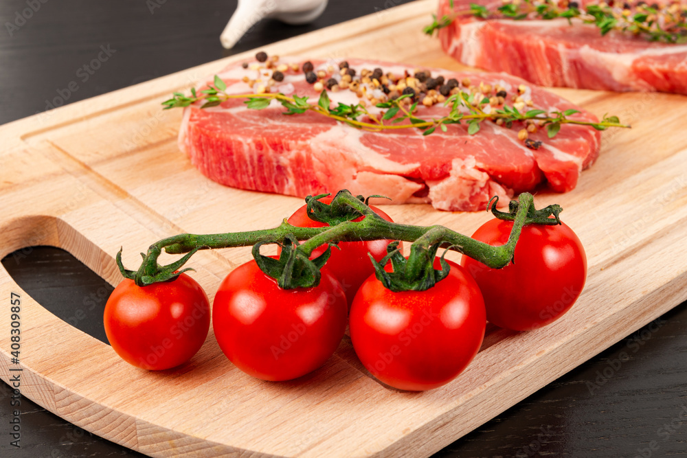 fresh chopped raw pork steaks with seasoning, ripe tomatoes and thyme on a cutting kitchen board on a black wooden table fresh chopped raw pork steaks with seasoning, ripe tomatoes and thyme on a cutting kitchen board on a black wooden table