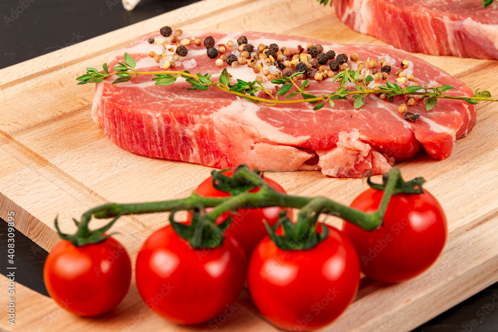fresh chopped raw pork steaks with seasoning, ripe tomatoes and thyme on a cutting kitchen board on a black wooden table fresh chopped raw pork steaks with seasoning, ripe tomatoes and thyme on a cutting kitchen board on a black wooden table