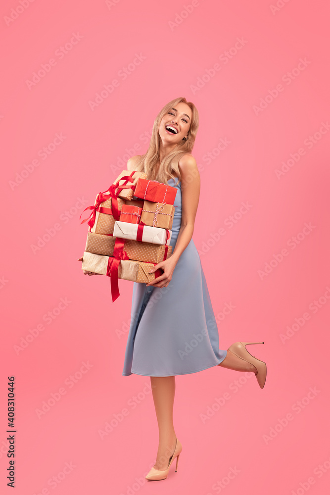 Full length portrait of beautiful blonde in dress holding heap of gift boxes on pink studio background Full length portrait of beautiful blonde in dress holding heap of gift boxes on pink studio background
