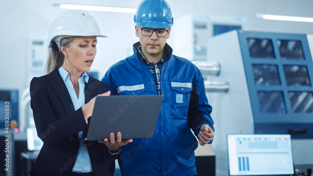 Female Manager and Project Engineer Wearing Hardhats Use Laptop in Industrial Factory, Talk, Plan Productivity Optimization. Production Line Workers Operate CNC Machinery, Program robot arm Female Manager and Project Engineer Wearing Hardhats Use Laptop in Industrial Factory, Talk, Plan Productivity Optimization. Production Line Workers Operate CNC Machinery, Program robot arm