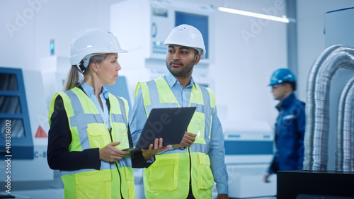 Chief Engineer and Project Manager Wearing Safety Vests and Hard Hats Walk Through Modern Factory, Talking, Optimizing Production Line. Industrial Facility: Professionals Working on Machinery