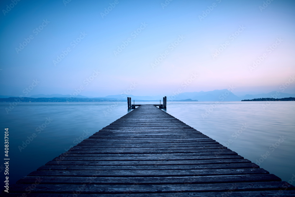 Fototapeta premium An old wooden pier extends into the clear blue waters of the lake. A solitary path towards the calm and peaceful silence of nature, with the sweet sound of the waves.