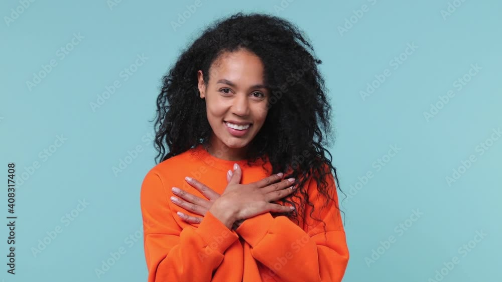 Smiling good kind young african american woman curly hair 20s years old wearing orange sweatshirt ask who me oh it so sweet put hands on chest isolated on pastel blue color background studio portrait Smiling good kind young african american woman curly hair 20s years old wearing orange sweatshirt ask who me oh it so sweet put hands on chest isolated on pastel blue color background studio portrait