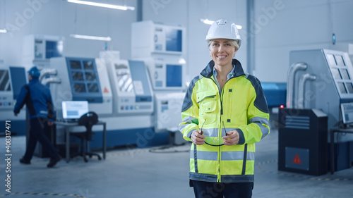 Beautiful Smiling Female Engineer Wearing Safety Vest and Hardhat Holds Safety Goggles. Professional Woman Working in the Modern Manufacturing Factory. Facility with CNC Machinery and robot arm © Gorodenkoff