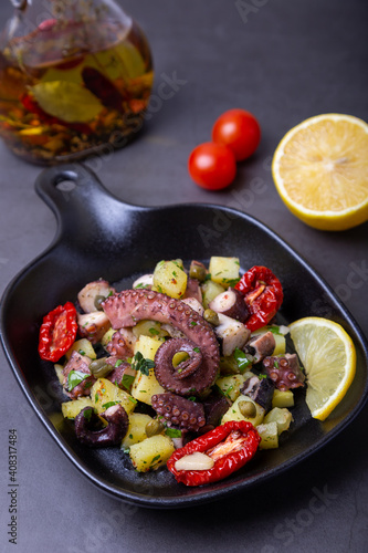 Warm salad with octopus, potatoes, sun-dried tomatoes, capers and lemon in a black small pan. Close-up, black background.