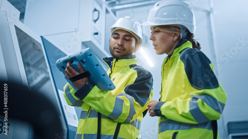 Chief Engineer and Project Manager Wearing Safety Vests and Hard Hats, Use Digital Tablet Controller in Modern Factory, Talking, Programming Machine For Productivity. Low Angle Portraits