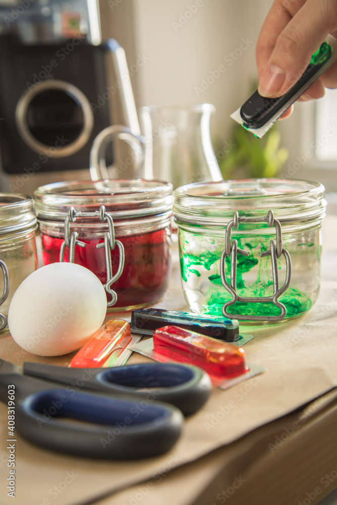 Frau gießt flüssige Ostereier Kaltfarbe Grün in Glas mit Essig Wasser auf Papier Unterlage in Küche neben Orange, Gelb, Blau, Rot in Verpackung, gekochte Eier und Schere zum Färben zu Ostern Frau gießt flüssige Ostereier Kaltfarbe Grün in Glas mit Essig Wasser auf Papier Unterlage in Küche neben Orange, Gelb, Blau, Rot in Verpackung, gekochte Eier und Schere zum Färben zu Ostern