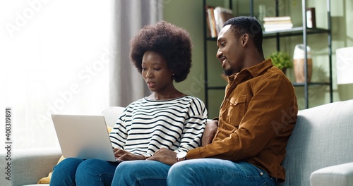 Young african american couple sitting at home on sofa and looking at laptop. Husband observes how wife works remotely.