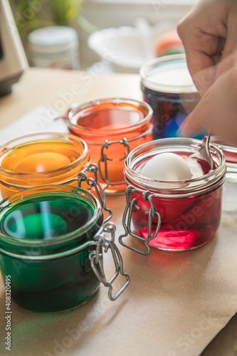 Frau taucht gekochte Eier mit Löffel in bunte Ostereier Kaltfarben in Rot, Orange, Gelb, Blau, Grün in Glas auf Papier Unterlage in Küche zum Färben zu Ostern