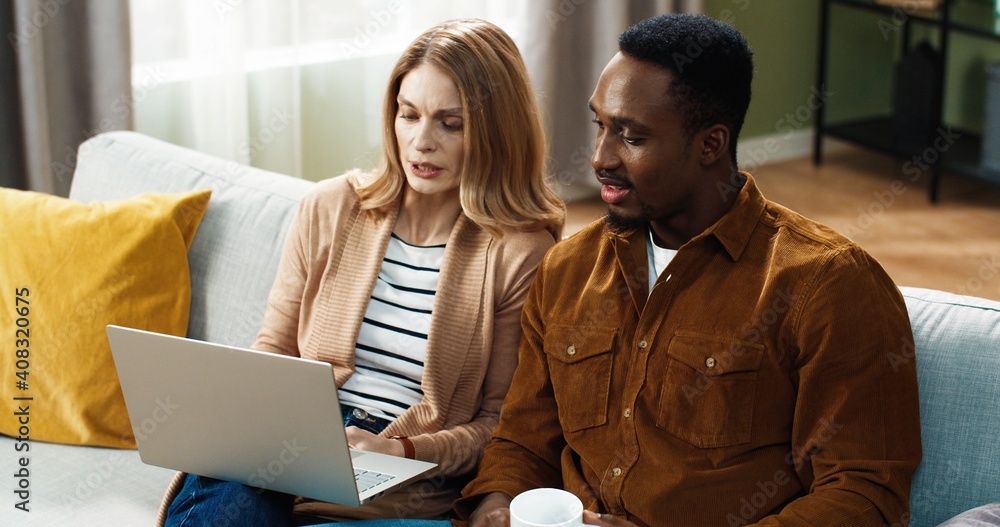 Beautiful multinational couple sitting at home on sofa and spending leisure time together. Beautiful caucasian woman working on laptop, african american man drinking hot tea. Beautiful multinational couple sitting at home on sofa and spending leisure time together. Beautiful caucasian woman working on laptop, african american man drinking hot tea.