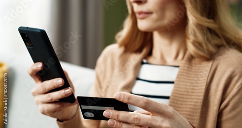 Close up of happy woman holding smartphone and bank card in hands and rejoicing in successful shopping and good news while sitting at home.