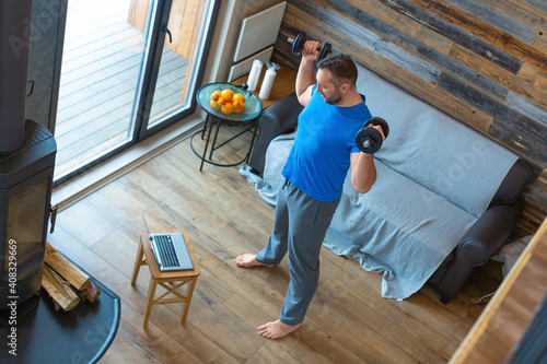A man works out at home with dumbbells in front of a laptop. He uses a tutorial from the internet.