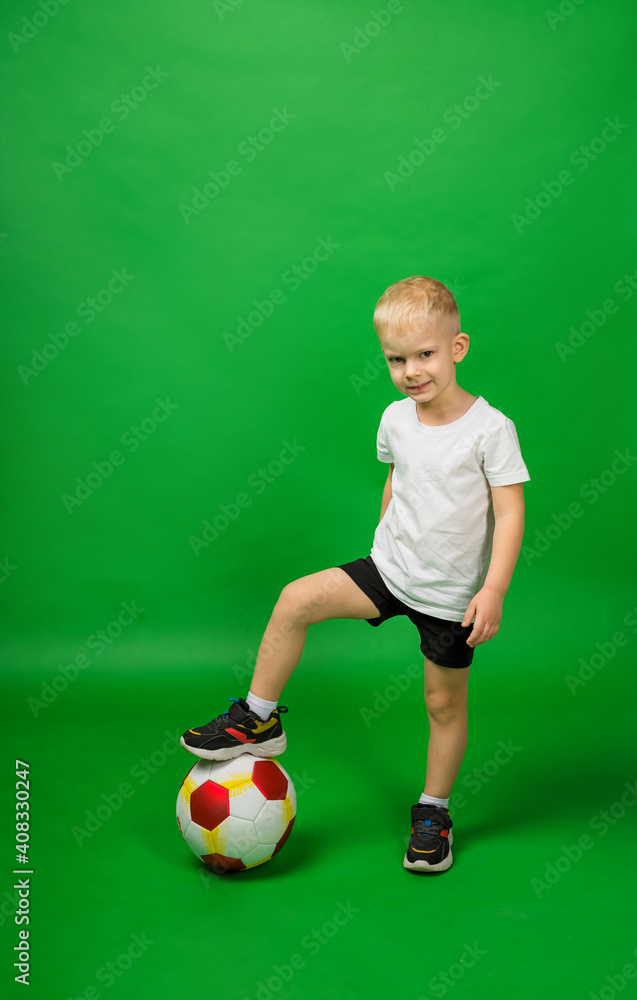 a little boy football player in a white T shirt and shorts holds the ball with his foot on a green background in full growth a little boy football player in a white T shirt and shorts holds the ball with his foot on a green background in full growth