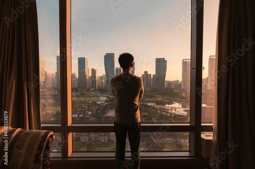 Young asian man standing and looking through window in a hotel. Sunrise over modern buildings in Songdo central park