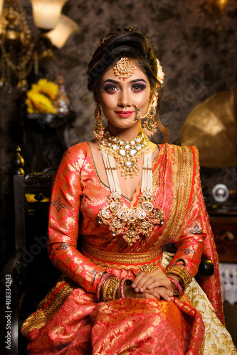 Leinwand Poster Magnificent young Indian bride in luxurious bridal costume with makeup and heavy jewellery sitting in a chair with classic vintage interior in studio lighting