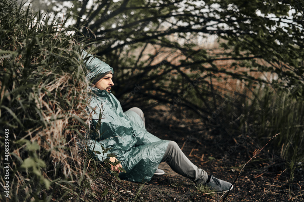 A man in a green raincoat sits on the ground in nature. A place for relaxation and rest.