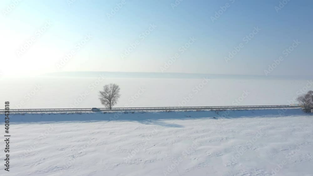 Aerial shot of white car riding through snow covered road near a frozen lake. SUV driving at countryside route near icy pond at sunny winter day. Flying over the auto moving through scenic landscape Aerial shot of white car riding through snow covered road near a frozen lake. SUV driving at countryside route near icy pond at sunny winter day. Flying over the auto moving through scenic landscape