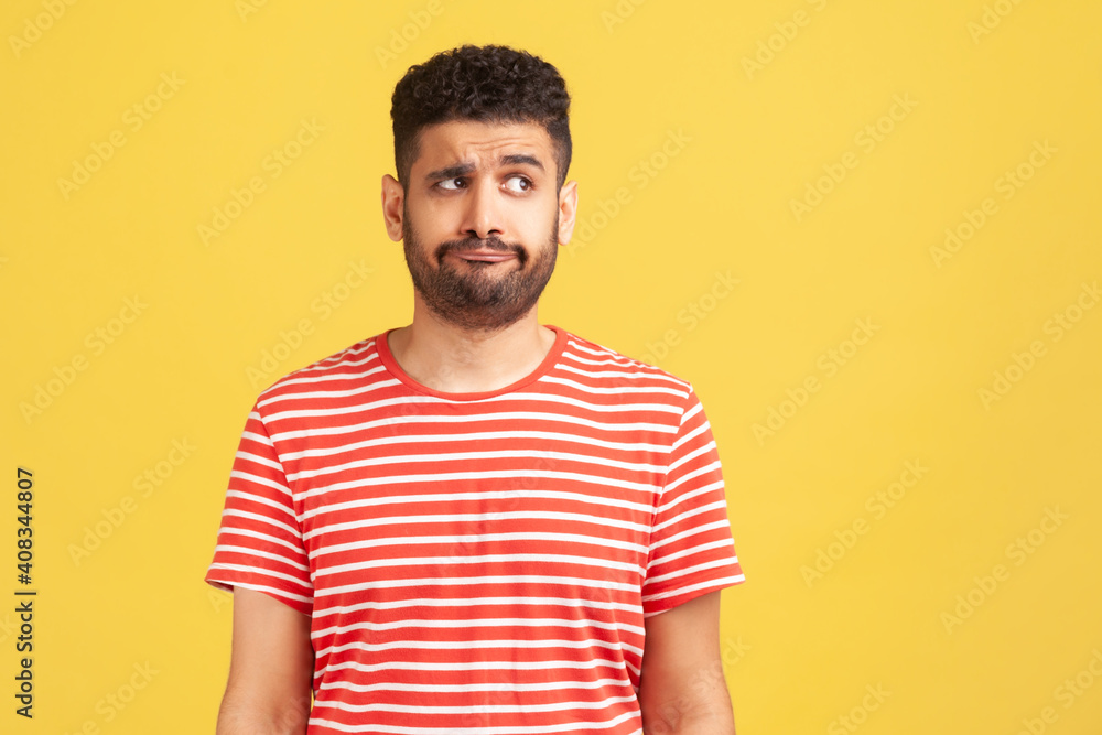 Fototapeta premium Doubtful bearded man in striped t-shirt standing with thoughtful expression, remembering or planning, thinking over answers