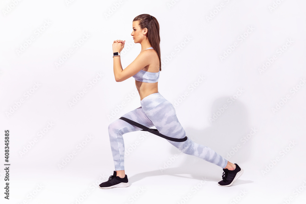 Side view concentrated athletic woman in white top and tights doing lunge with elastic latex resistance band on legs, pumping up muscles