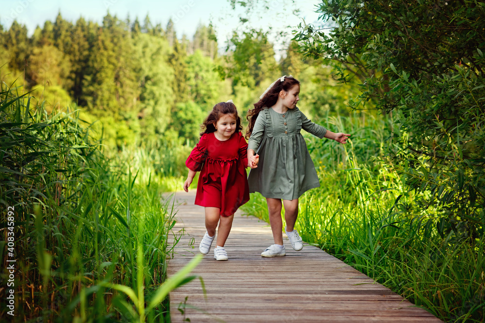 Fototapeta premium Two little girls with long hair in a green and red dress run along a wooden path in the Park. A fun childhood of the two sisters.