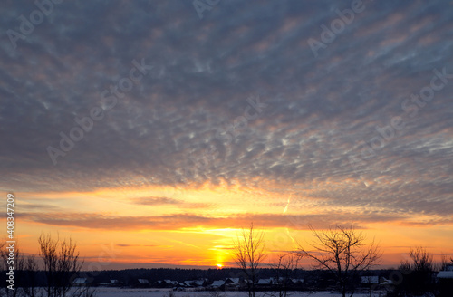 Golden rays of the sun in the sky with cumulus clouds over the village on a winter evening, rural landscape