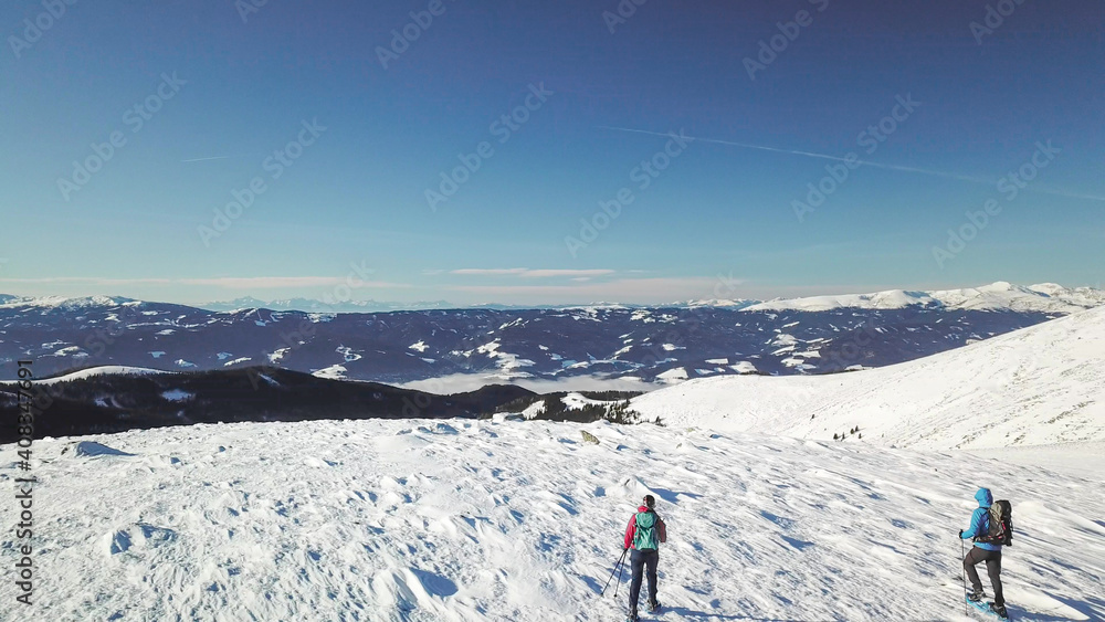 A drone shot of a couple wearing snow shoes hiking up to the peak of Speikkogel in Austrian Alps. The whole slope is covered with snow. many mountain chains in the back. Winter outdoor activity. Fun