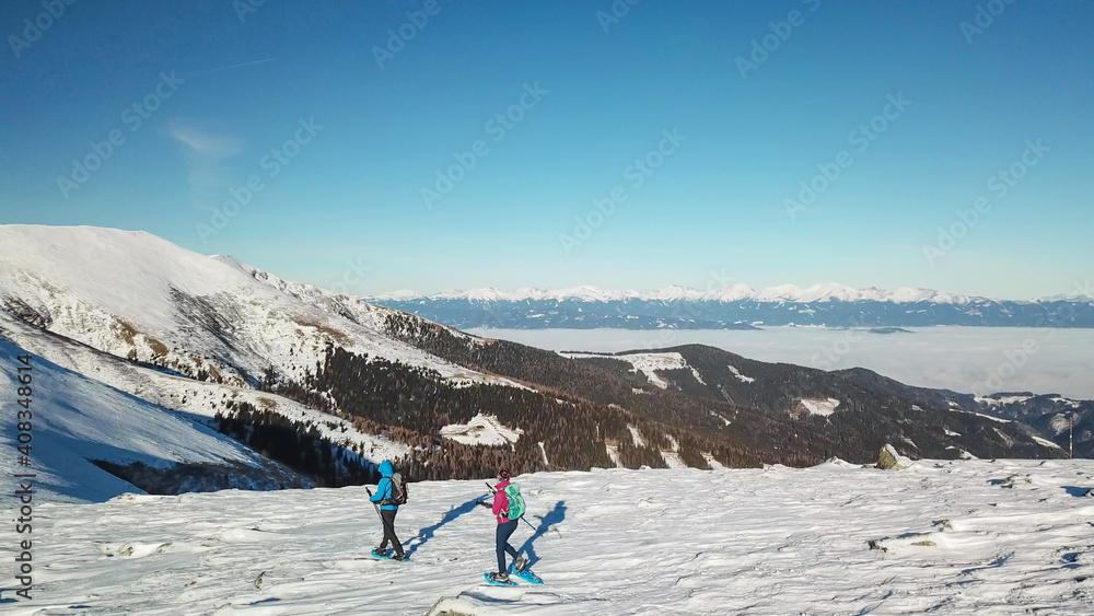 A drone shot of a couple wearing snow shoes hiking up to Amerinkogel's peak in Austrian Alps. Fresh powder snow. Many mountain chains in the back, valley shrouded in fog. Winter outdoor activity. Fun