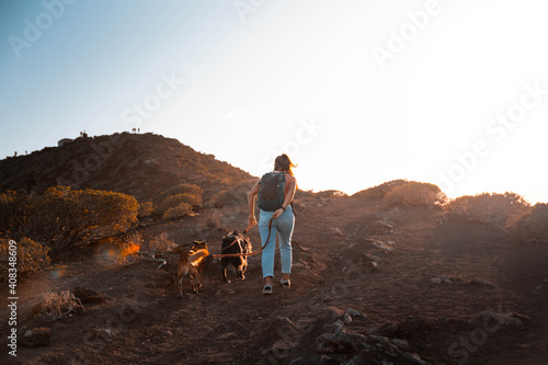 Woman from behind walking two dogs on a leash in the red mountain of Tenerife at sunset