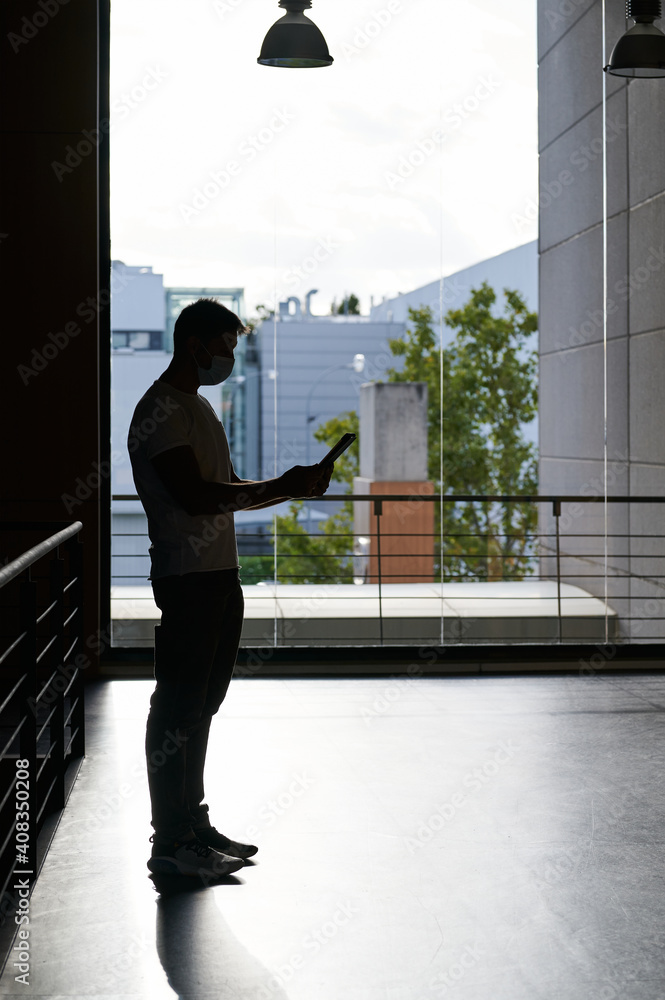 Fototapeta premium middle-aged man wearing a mask and using a tablet next to a large window. Natural light