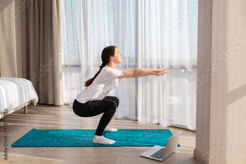 A young slender woman in sportswear trains at home for online lessons. There's a laptop on the floor. Side view. Concept of home sports training on videocommunication