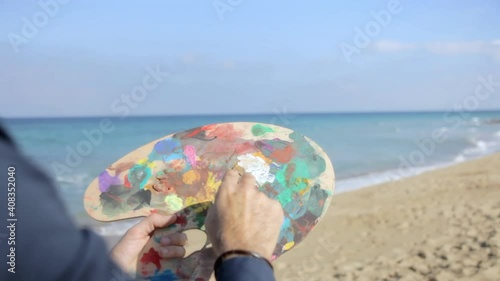 An inspired man (an artist), at a sandy beach, painting the sky with colors from a painter's palette. Creativity, inspiration. Handheld shot.
 photo