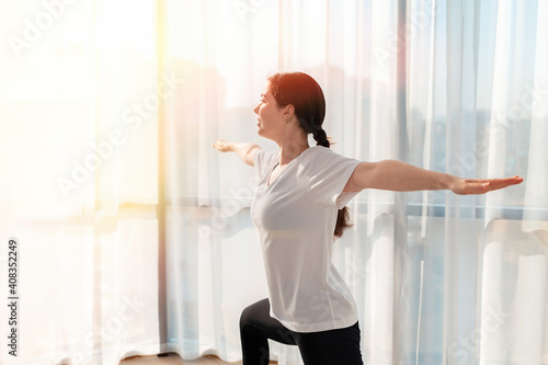 Yoga. A young woman trains at home, and smiles while doing a warrior pose. Sunlight from the window. The concept of home workouts