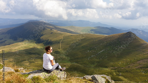 Woman in hiking outfit sitting at a big rock and enjoys the view, while hiking to Speikkogel, Austrian Alps. The mountains around are lush green, not so sharp. Sunny summer day. Serenity and calmness photo