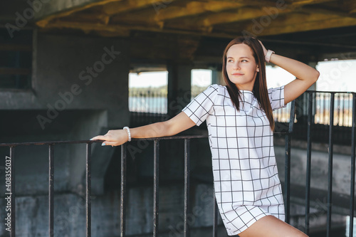 Pretty woman leaning on metal fence under bridge at sunny summer day. Urban style