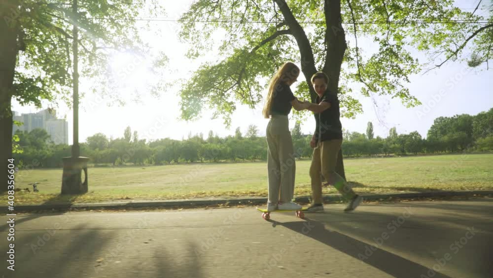 Wide shot of happy Caucasian teenage boy teaching girl riding skate in sunlight outdoors. Side view of cheerful boyfriend holding hand of girlfriend skateboarding in sunbeam in summer park. Wide shot of happy Caucasian teenage boy teaching girl riding skate in sunlight outdoors. Side view of cheerful boyfriend holding hand of girlfriend skateboarding in sunbeam in summer park.