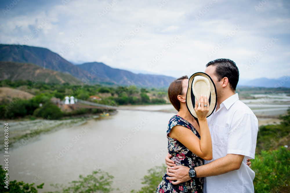 Two people in love kiss while covering their faces with a hat next to a river and mountains